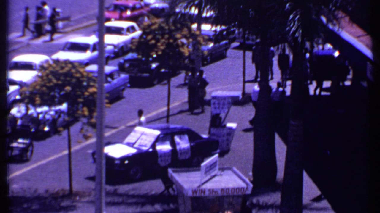 1969 Kenya balcony-level view of a palm-lined urban street with parked cars, pedestrians, road signs, and visible storefront signage.