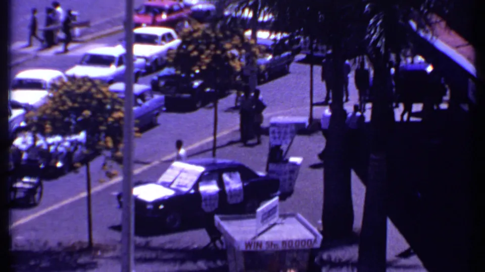 1969 Kenya balcony-level view of a palm-lined urban street with parked cars, pedestrians, road signs, and visible storefront signage.
