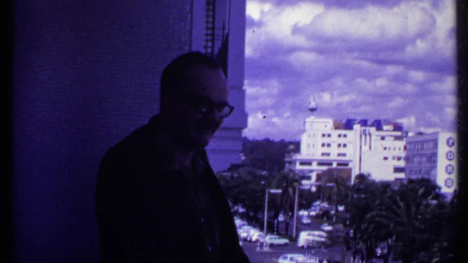 A middle-aged man in glasses stands on a balcony in 1969 Kenya, smiling slightly as a bright city street and building appear behind him.