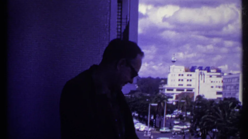 1969 Kenya home-movie frame showing a man in profile on a balcony, with traffic, palms, signs, and a distant urban building beyond.