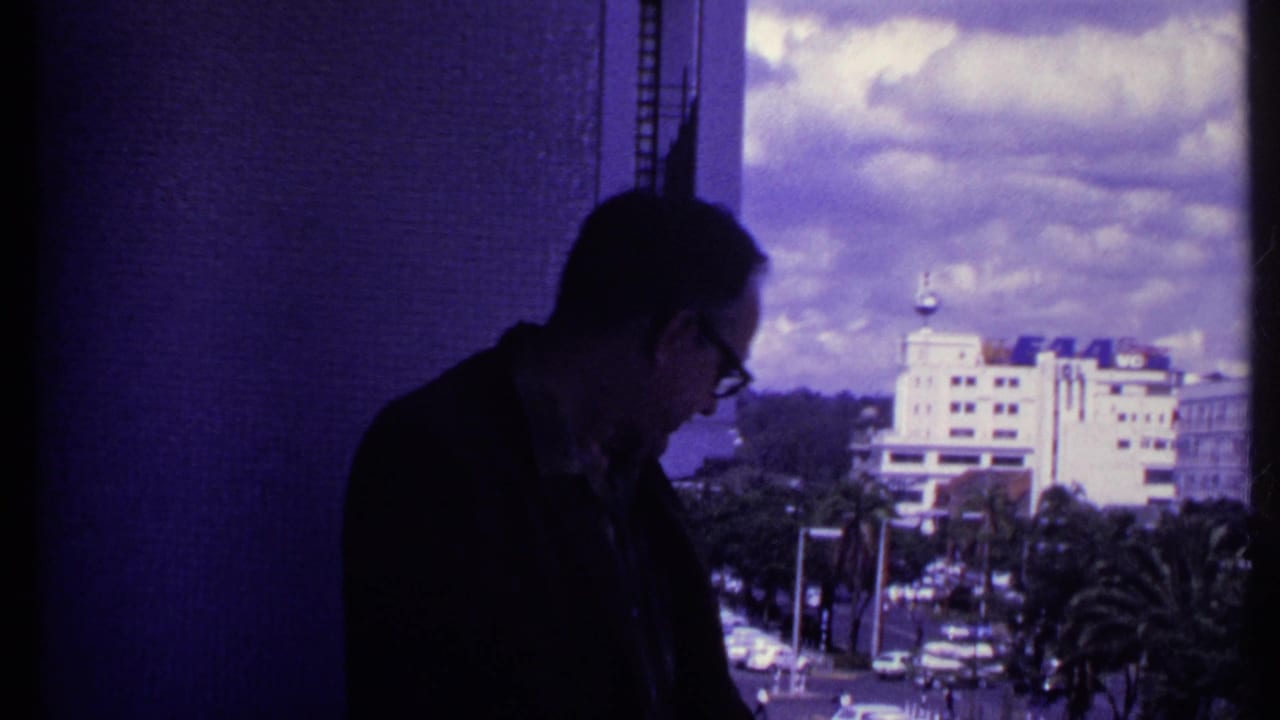 Man in glasses stands in shadow near a balcony view of a busy street and distant building.