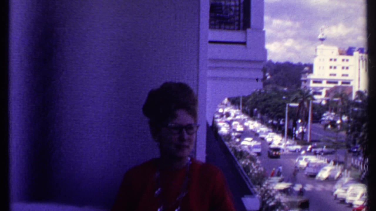 Woman in glasses and red top standing on a balcony above a busy city street.