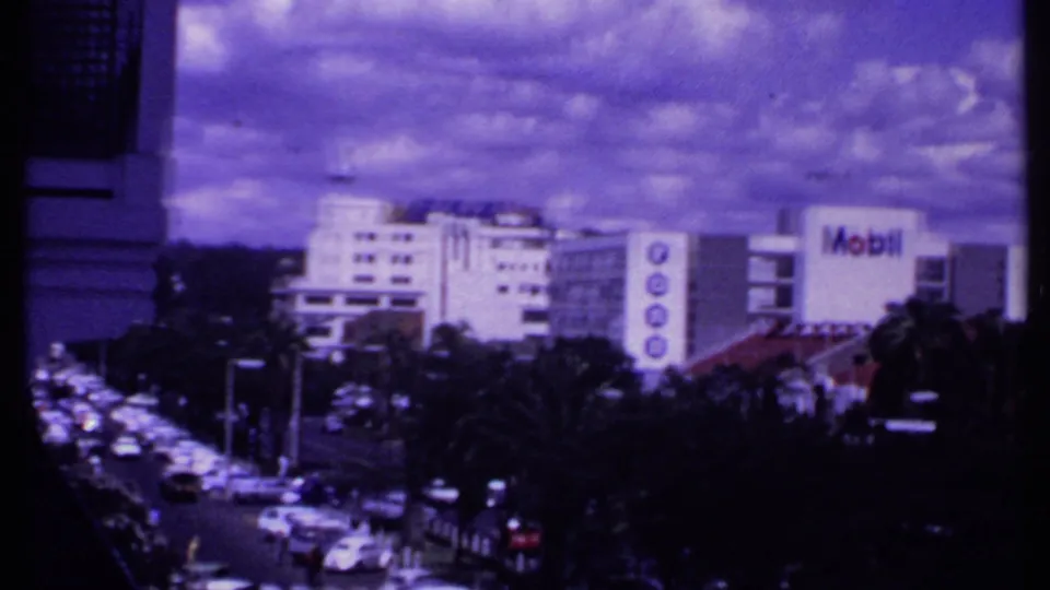 A panning 1969 Kenya street view shows dense traffic, palms, white commercial buildings, and a prominent Mobil sign.