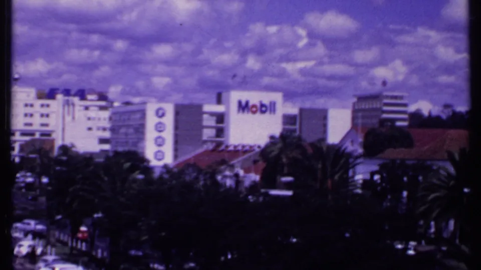 The pan ends on a wider 1969 Kenya city view with traffic below, palms in the foreground, and a Mobil sign on a building.