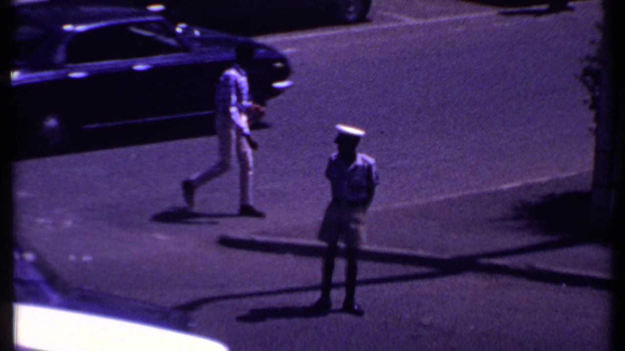 Uniformed man stands near a busy intersection with cars and pedestrians in the street.
