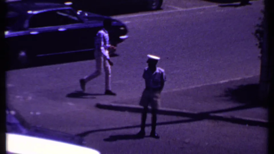 In 1969 Kenya footage, a uniformed man waits near a sunlit intersection as cars and pedestrians move through traffic.