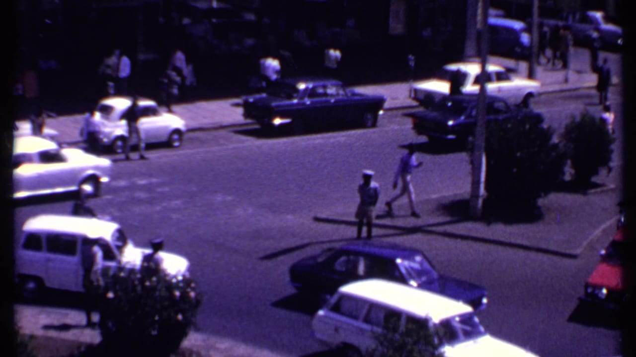 Cars pass through an intersection while pedestrians cross near uniformed figures.
