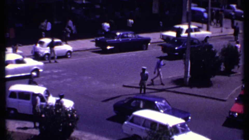 Elevated 1969 Kenya street view with pedestrians crossing between mid-century cars and uniformed personnel near the curb.