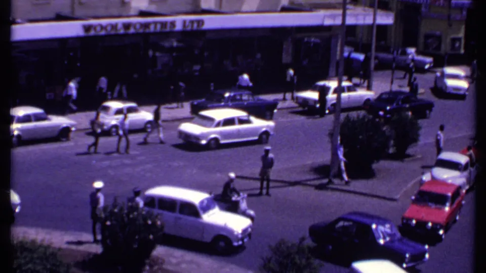 1969 Kenya city corner showing pedestrians, vintage cars, uniformed figures, and a Woolworths Ltd sign across the street.