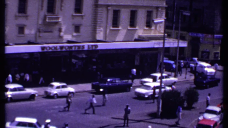 1969 Kenya urban street scene with the Woolworths Ltd storefront, moving cars, pedestrians, and a uniformed figure at the intersection.