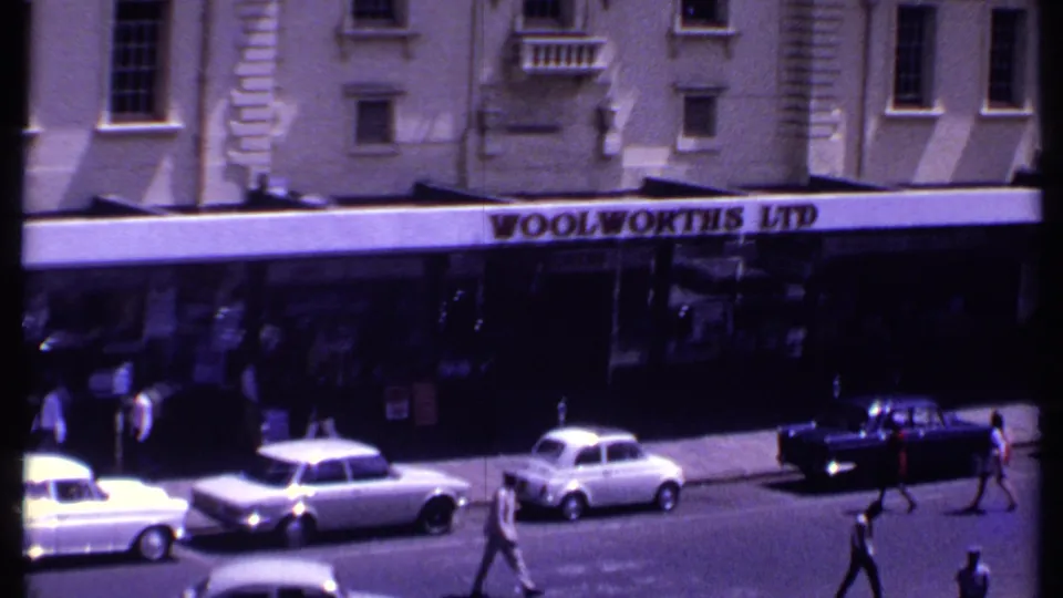 The camera has moved closer to the Woolworths Ltd sign, with sedans along the curb and pedestrians crossing the street in Kenya in 1969.