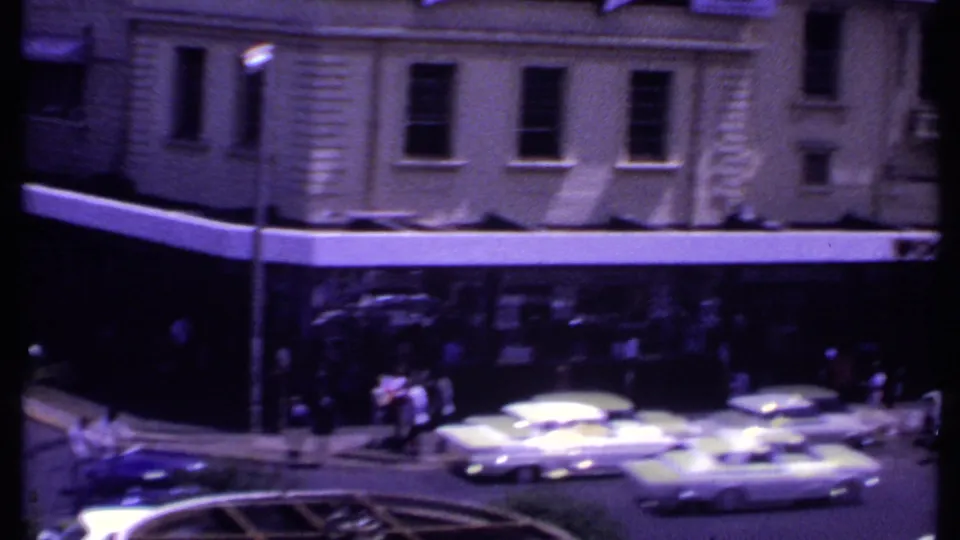 1969 Kenya urban street corner with pedestrians crossing near period cars and shopfronts beneath multi-story buildings.