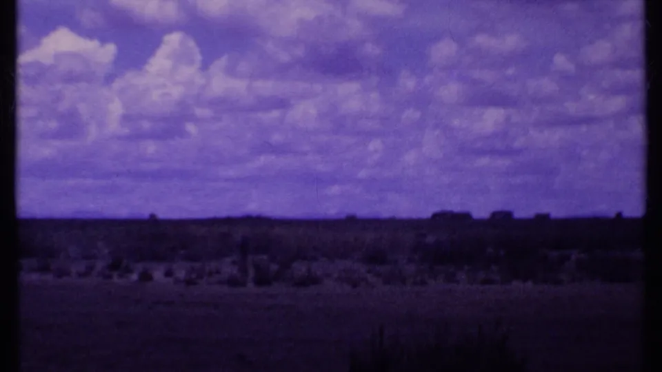 1969 Kenya 8mm frame with an open range, distant dark horizon forms, low shrubs, and a cloud-filled sky under a purple film cast.