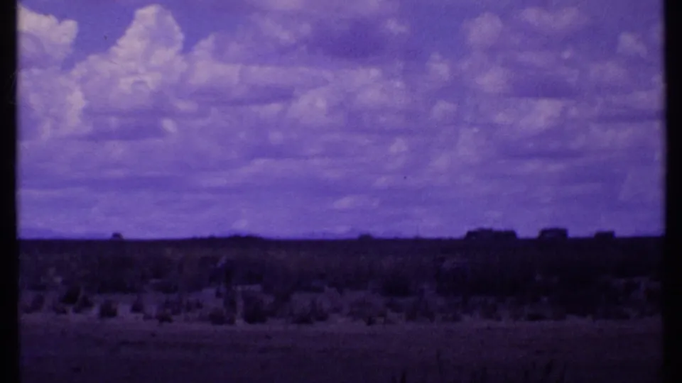 1969 Kenya 8mm frame showing a wide savanna horizon, clouded sky, scrubland, and distant wildlife forms.