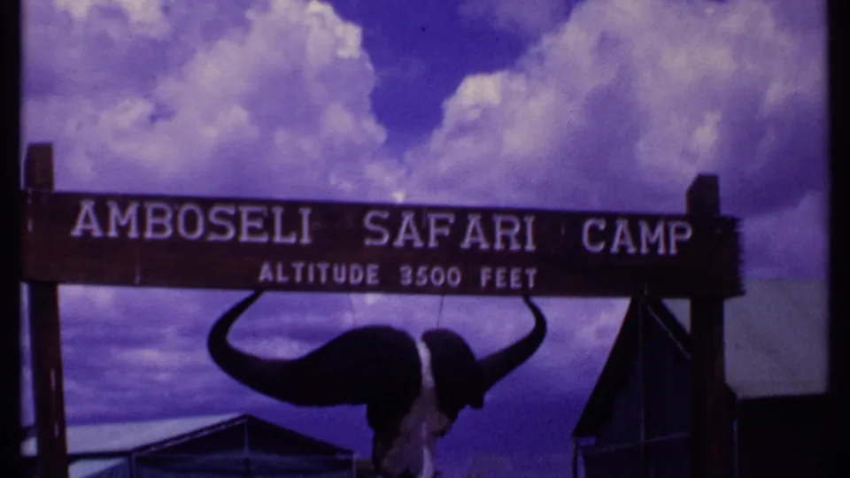 1969 Kenya footage frame with the Amboseli Safari Camp sign, altitude lettering, buffalo skull, dark posts, and dramatic clouds.