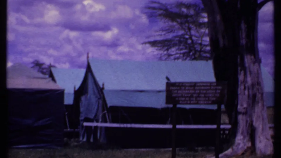 1969 Kenya visitor-shot frame showing canvas safari tents at Amboseli Safari Camp with trees, grass, and an information sign nearby.