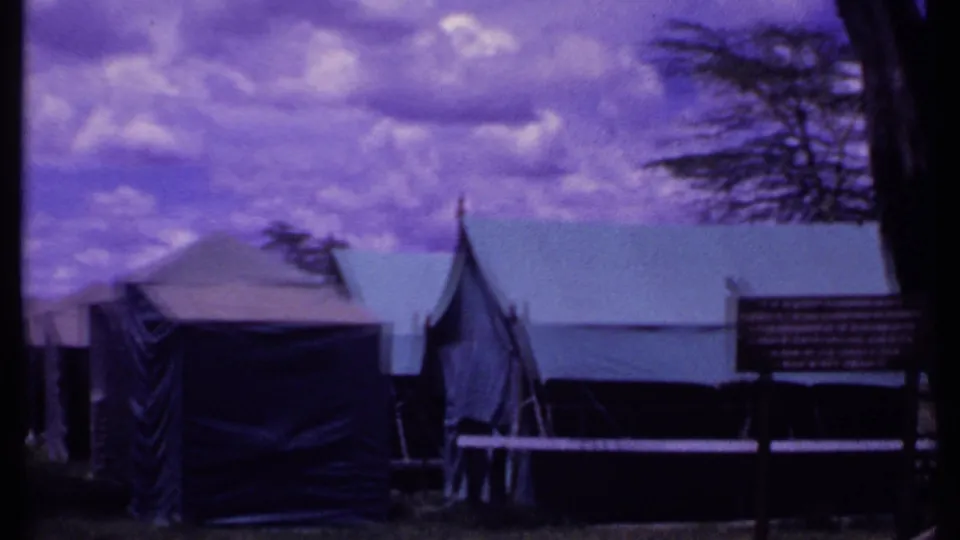 1969 Kenya home-movie frame of Amboseli Safari Camp grounds with canvas tents, trees, grass, and a small signboard under purple-shifted clouds.