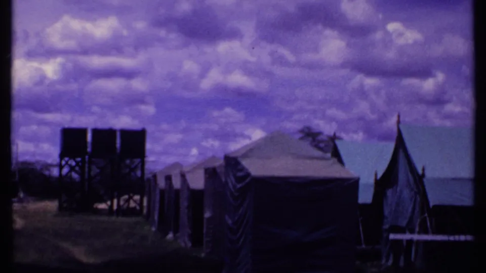 1969 Kenya home-movie frame with canvas tents, twin raised water tanks, fence posts, and open grassy camp ground.