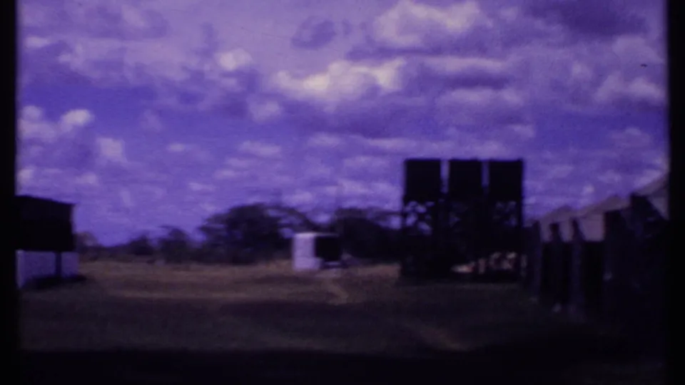 1969 Kenya 8mm frame looking across open grass toward elevated water tanks, distant tents, trees, and a purple-shifted sky.
