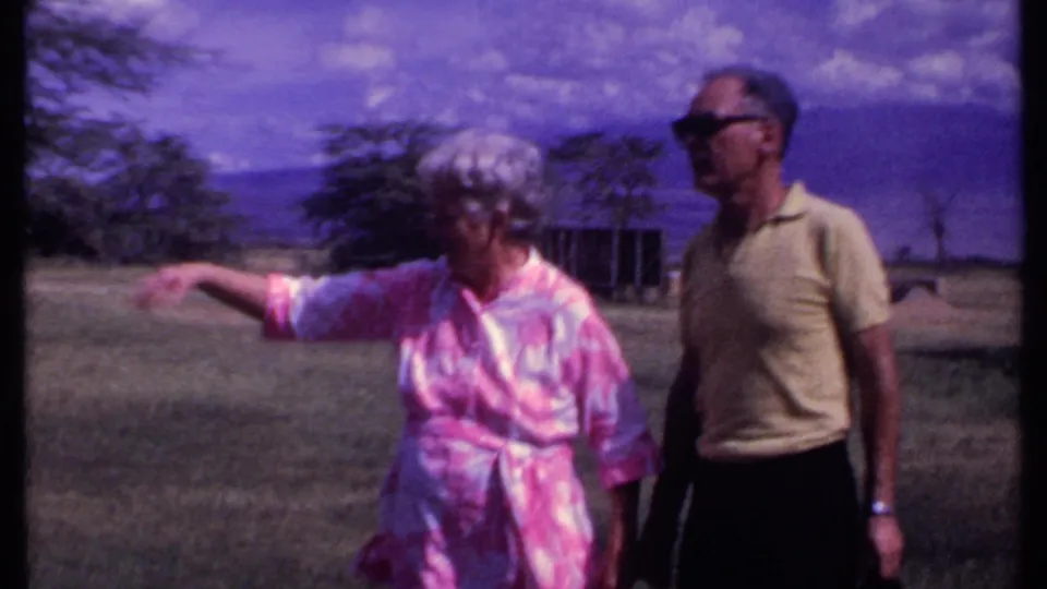 1969 Kenya home-movie frame of an older woman in a pink patterned dress waving beside an older man in sunglasses near camp structures.