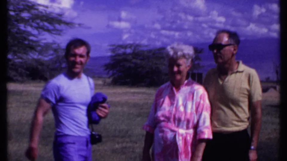 A younger man holding clothing or gear joins the older couple in the Kenya 1969 home-movie scene, all standing on open grass.