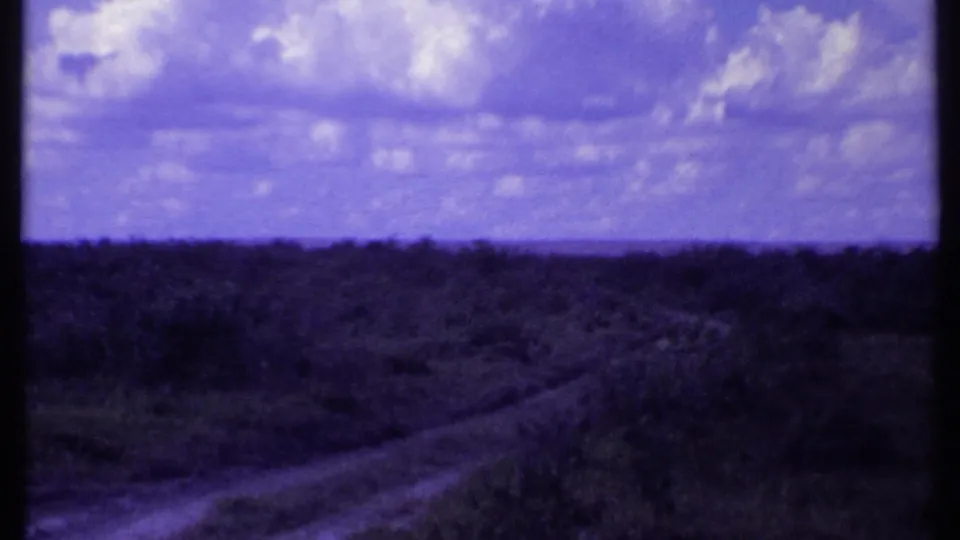 The 1969 Kenya 8mm frame shows open grassland and a faint dirt track receding toward a cloudy horizon.