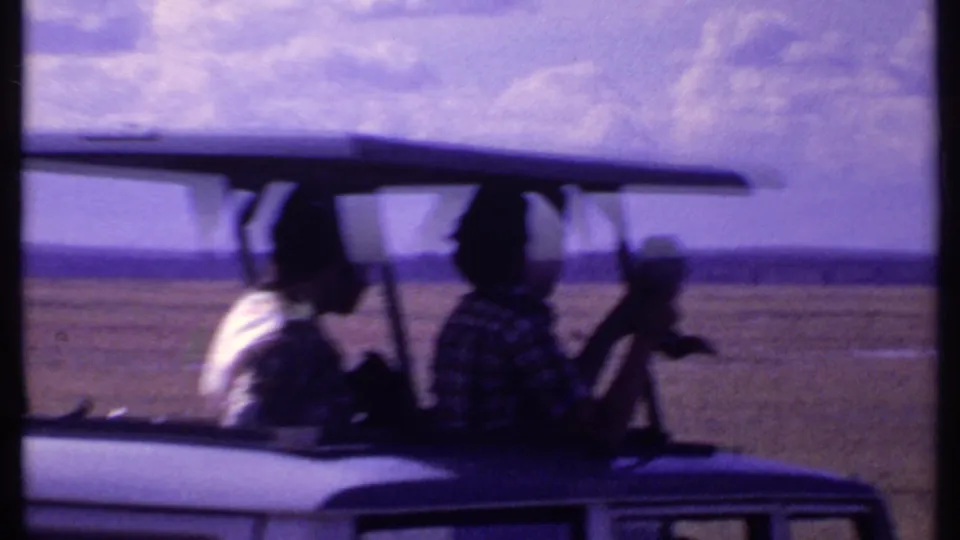 Kenya 1969 safari vehicle with seated tourists silhouetted beneath a roof canopy, crossing open scrubland.