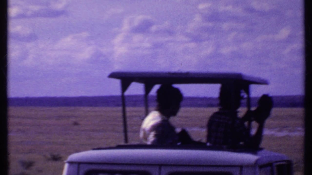 Two passengers ride in the back of an open-top safari jeep on a flat savanna.