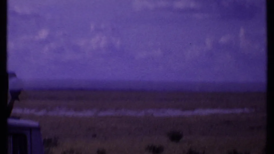 A 1969 Kenya safari vehicle appears at the edge of the frame as the camera looks across open grassland and a distant horizon.