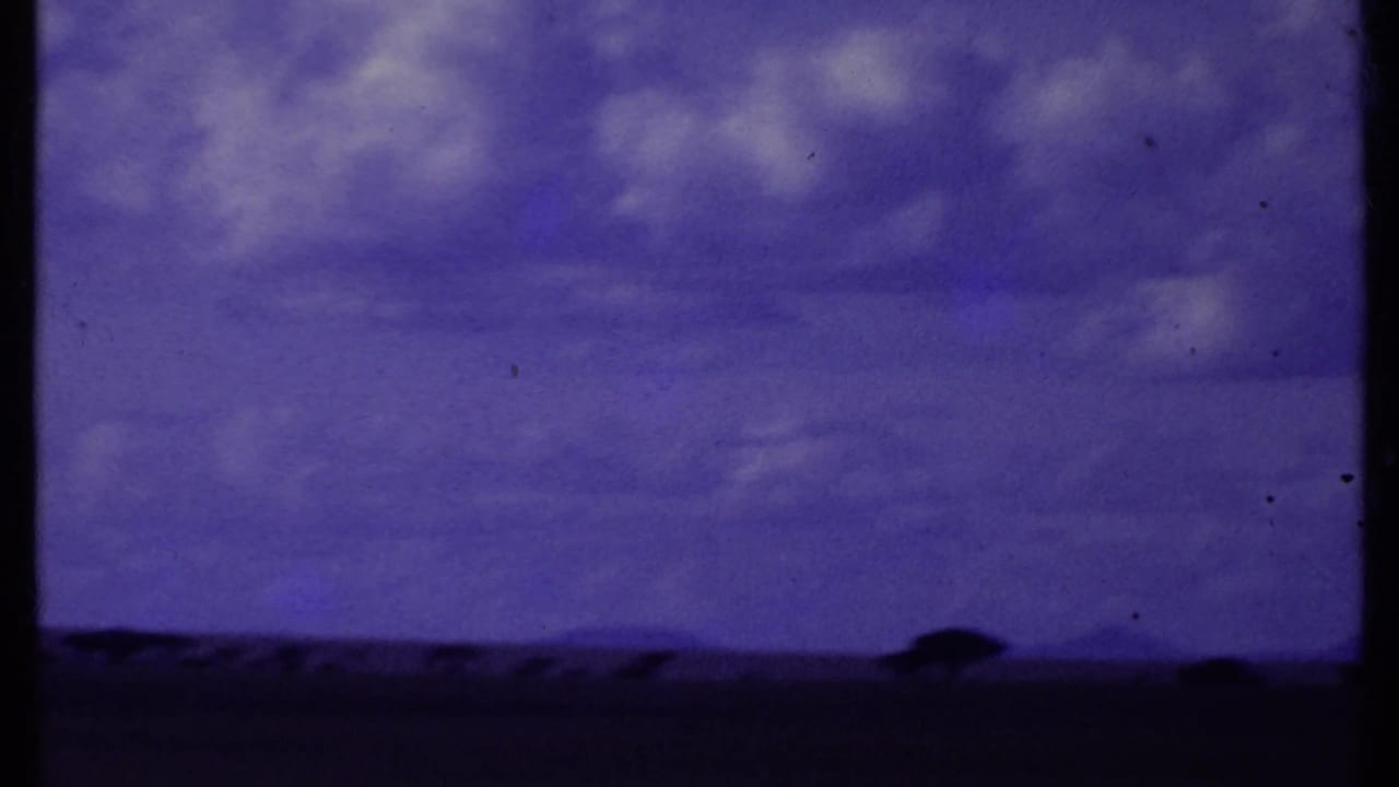 Kenya 1969 home-movie frame with a dark savanna horizon, scattered tree silhouettes, and a purple-blue sky with soft clouds.