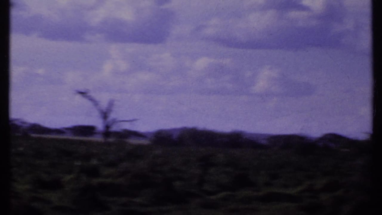 1969 Kenya 8mm frame after the elephant leaves view, showing open savanna, scattered trees, distant hills, and a clouded sky.