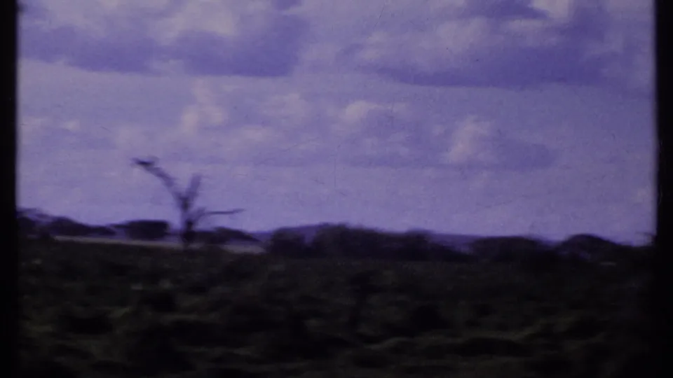 1969 Kenya 8mm frame after the elephant leaves view, showing open savanna, scattered trees, distant hills, and a clouded sky.