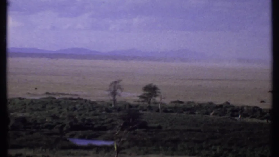 1969 Kenya 8mm frame looking across open savanna, scattered trees, a small waterhole, and hazy distant hills.