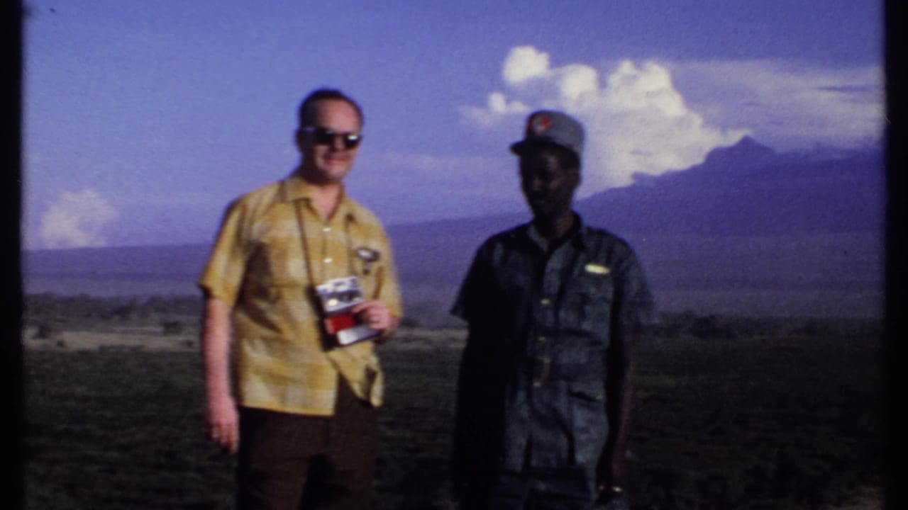In Kenya 1969 home-movie footage, a tourist with a camera stands beside a local uniformed man above a wide valley.