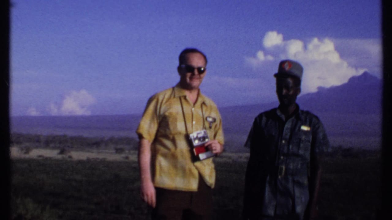 A man with a camera stands beside a uniformed man against a distant mountain view.