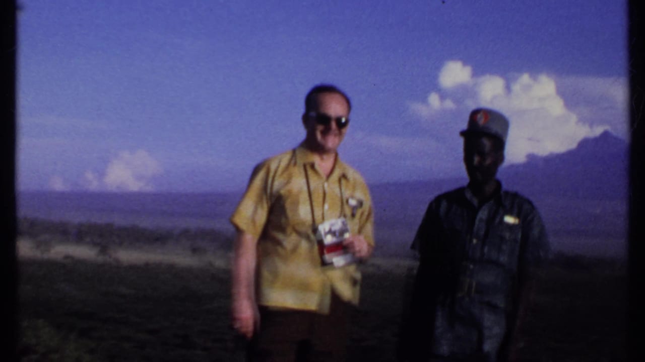 A handheld 8mm frame from Kenya in 1969 shows two men at a valley overlook, one holding a camera and the other in a dark uniform.