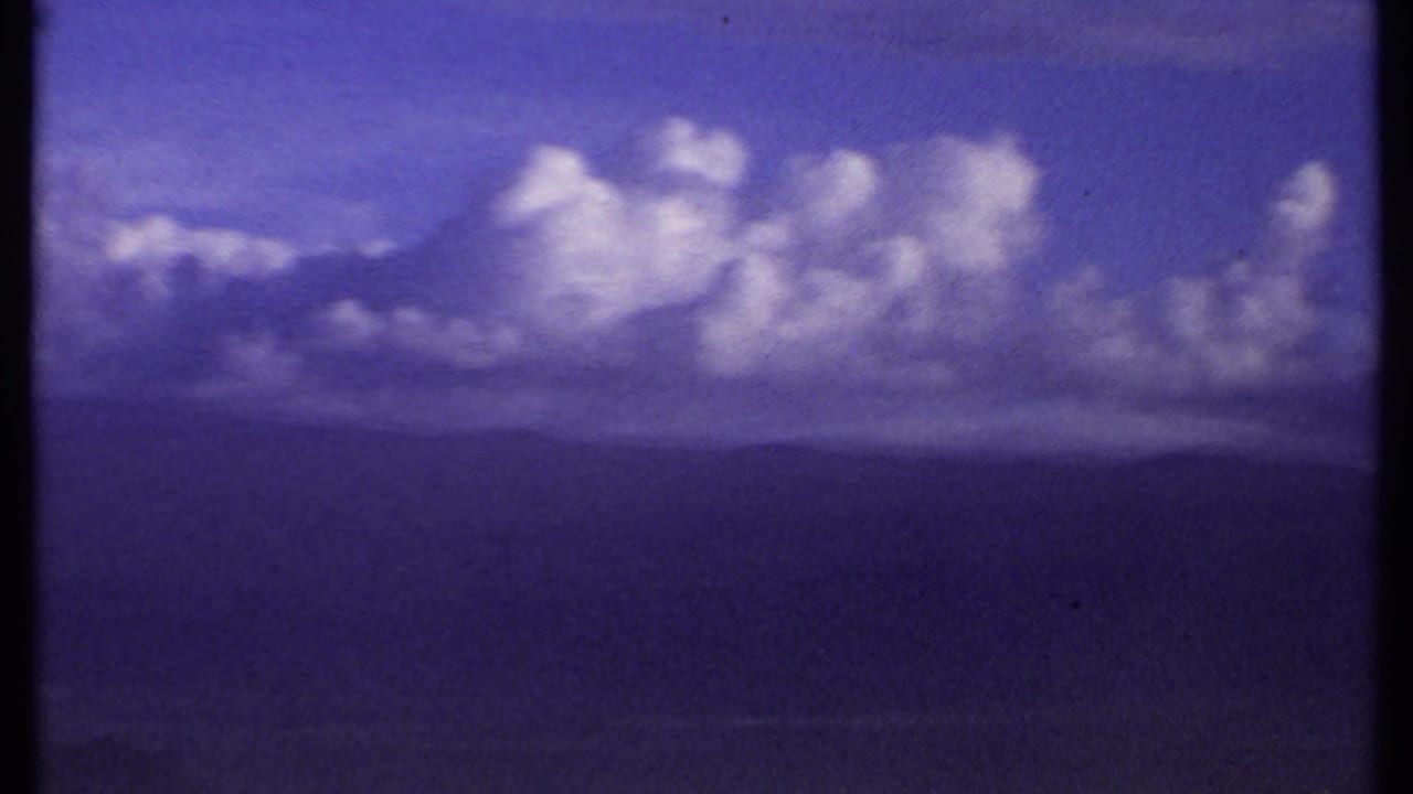 1969 Kenya 8mm frame showing brighter cumulus clouds drifting over a low, hazy mountain horizon.