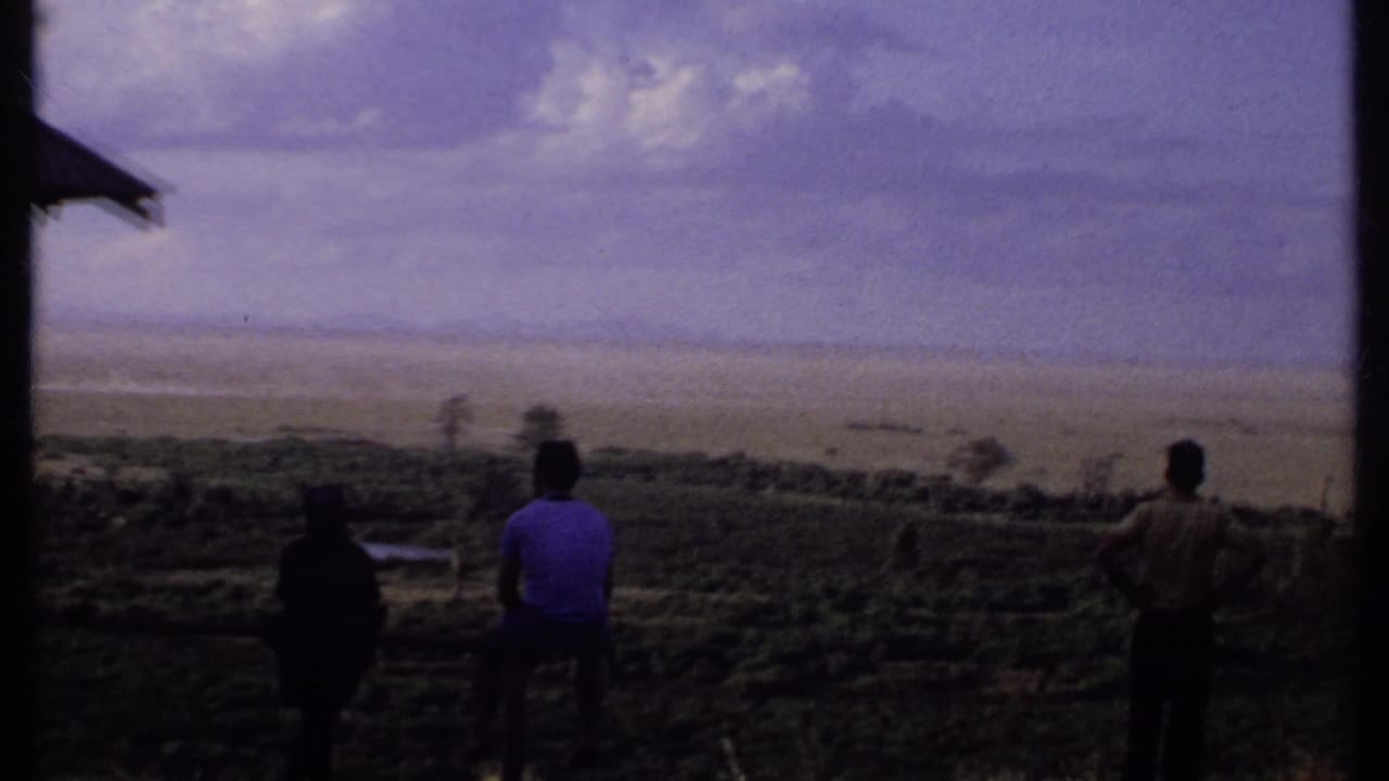 1969 Kenya 8mm frame showing three people from behind on a hillside, with a partial roof edge and broad landscape beyond.