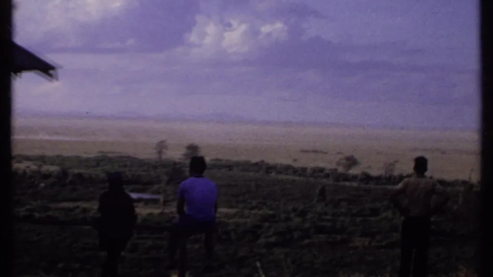 1969 Kenya 8mm frame showing three people from behind on a hillside, with a partial roof edge and broad landscape beyond.