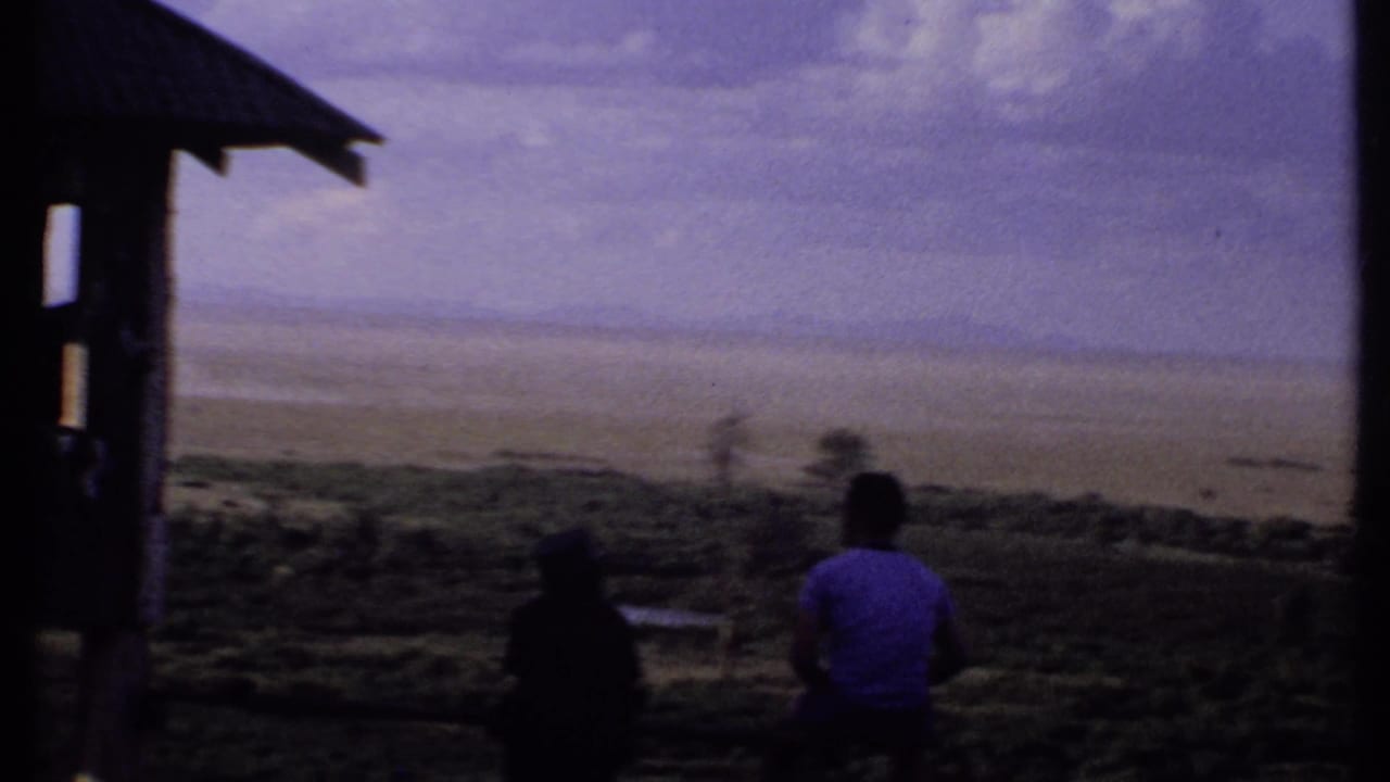 1969 Kenya 8mm frame with two silhouetted people near a shelter post, looking across a wide pale plain under clouded sky.