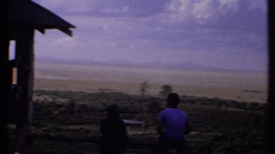 1969 Kenya 8mm frame with two silhouetted people near a shelter post, looking across a wide pale plain under clouded sky.