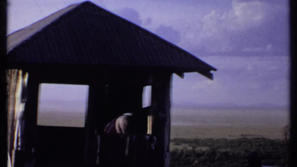 1969 Kenya 8mm view of a rustic shelter with a person leaning near the doorway and a hazy plain beyond.