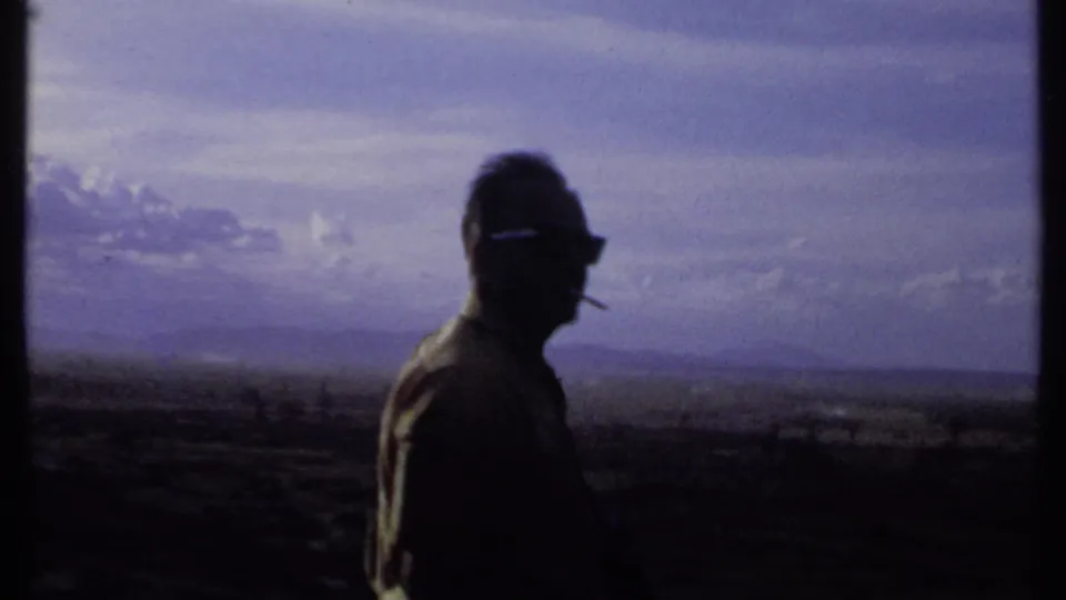 The man turns in profile in this 1969 Kenya home-movie frame, with sunglasses and a cigarette visible against the distant landscape.