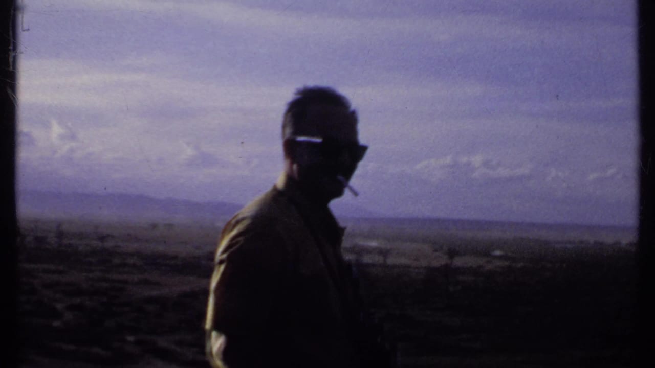 A silhouetted man in sunglasses and a collared shirt holds a cigarette while standing before a hazy Kenyan landscape in 1969 footage.