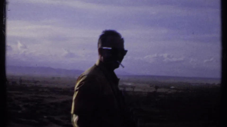 A silhouetted man in sunglasses and a collared shirt holds a cigarette while standing before a hazy Kenyan landscape in 1969 footage.