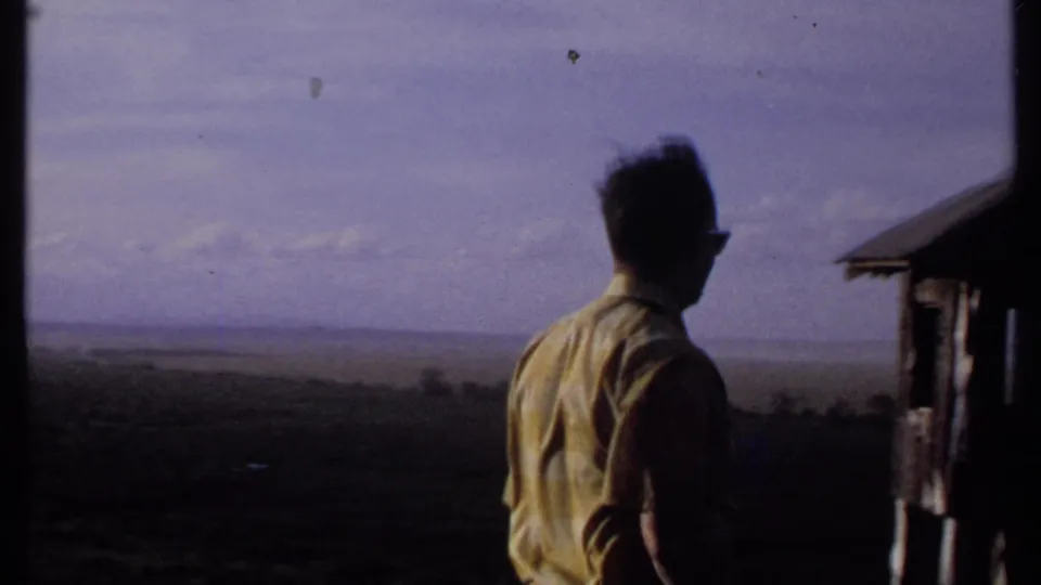 In 1969 Kenya home-movie footage, a man in a yellow shirt stands near a small shelter and looks across a broad distant landscape.