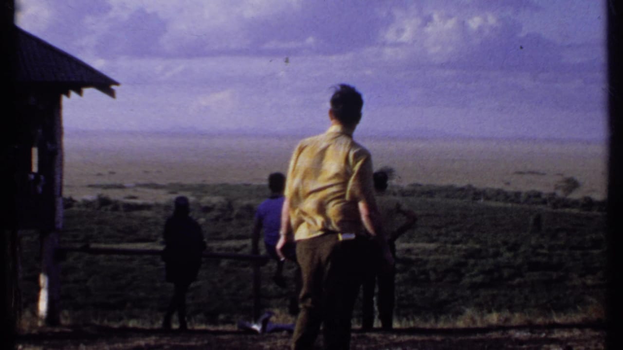 A man in a yellow shirt stands with other visitors at a Kenya overlook, with a small structure at left and distant terrain beyond.