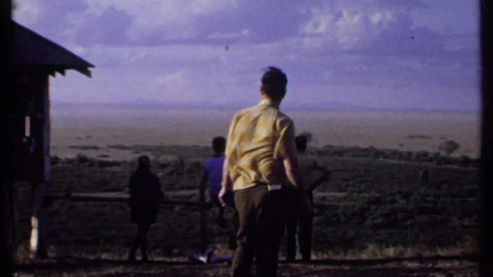 A man in a yellow shirt stands with other visitors at a Kenya overlook, with a small structure at left and distant terrain beyond.
