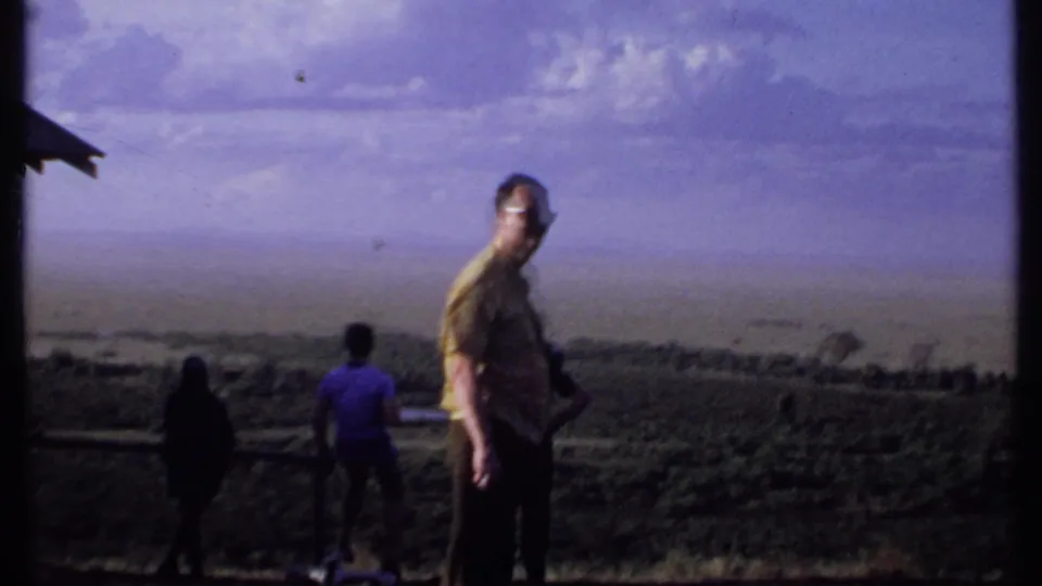 1969 Kenya 8mm frame of adult men at a grassy overlook, one in a yellow shirt and sunglasses facing the camera.