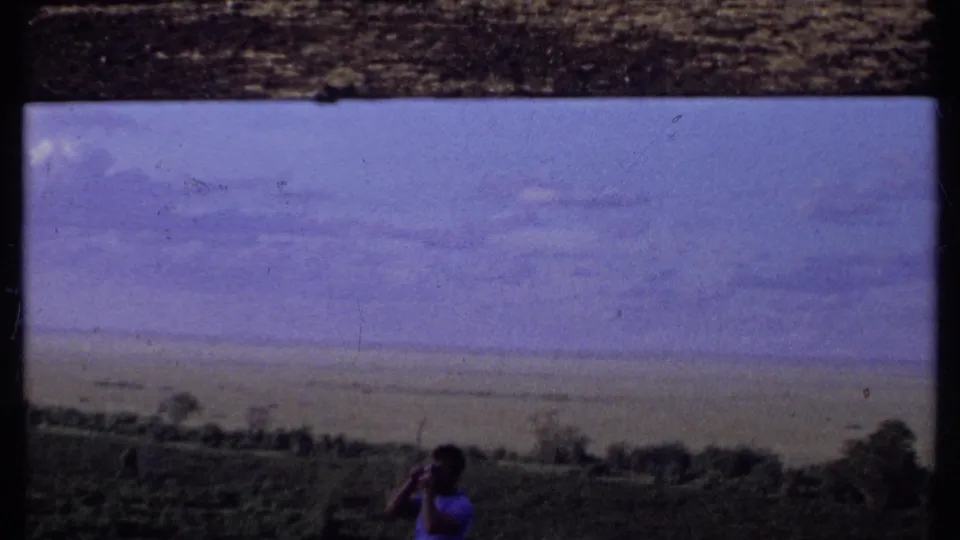 1969 Kenya travel frame showing a man holding a camera toward the open savanna under a blue-purple sky.